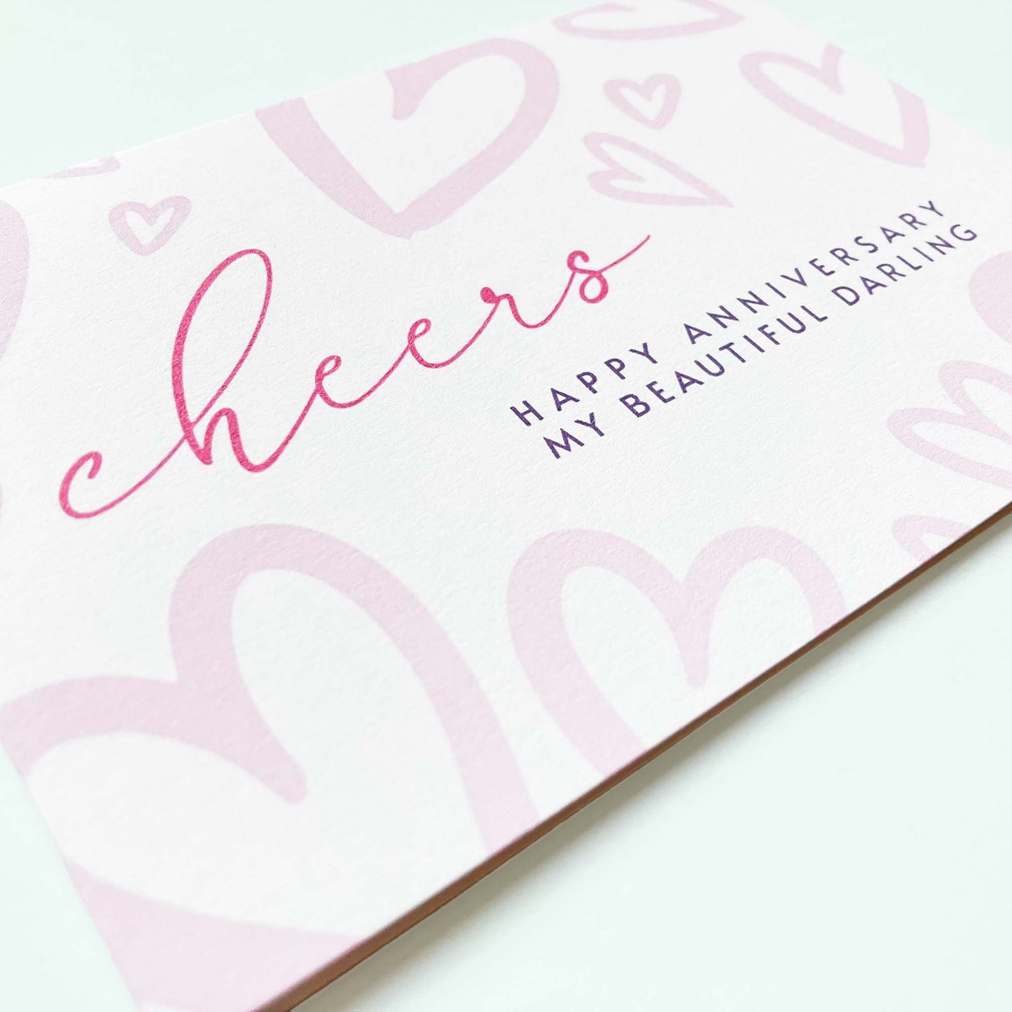 Close-up angle shot of Anniversary card with 'cheers' and 'Happy Anniversary My Beautiful Darling' text on a white background and heart illustration pattern.