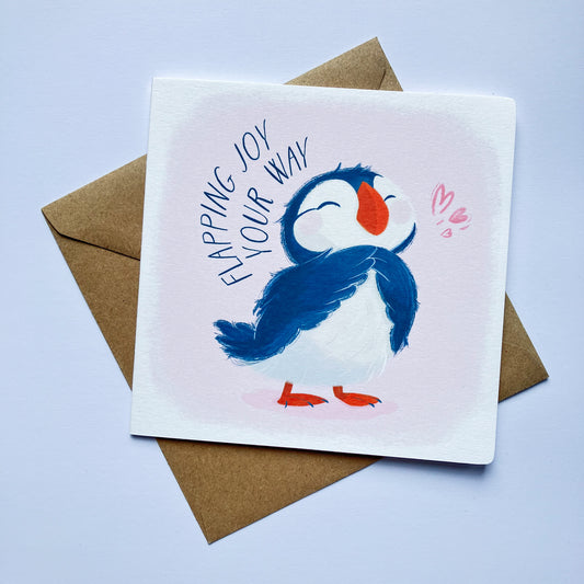 A square birthday card with an illustration of a baby puffin, placed on a kraft brown envelope.
