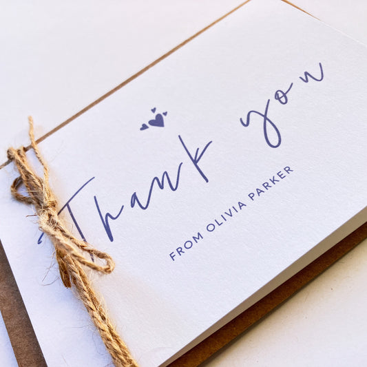 Angle view of a personalised thank you card with a handwritten "Thank you" text in the middle. On top of the text, there are four heart shapes, one big and three small ones on the left, right and top. Below the "Thank you" text is the personalisation with example one shown as "FROM OLIVIA PARKER". The card's background is soft lavender one and the text and illustrations are in dark muted purple. The card is wrapped on the left side with a natural baker thick thread.