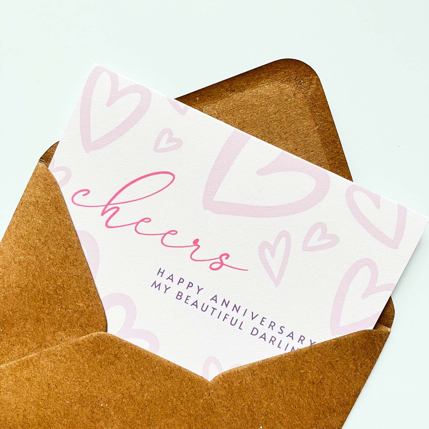Anniversary card with 'cheers' and 'Happy Anniversary My Beautiful Darling' text on a white background and heart illustration pattern in a Kraft brown envelope on a light green background.