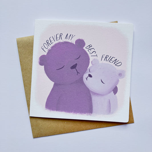 A greeting card with a whimsical illustration of two bears, one purple and one pink, with the text 'FOREVER MY BEST FRIEND' above them. The card is placed on a white background with a brown envelope behind it.