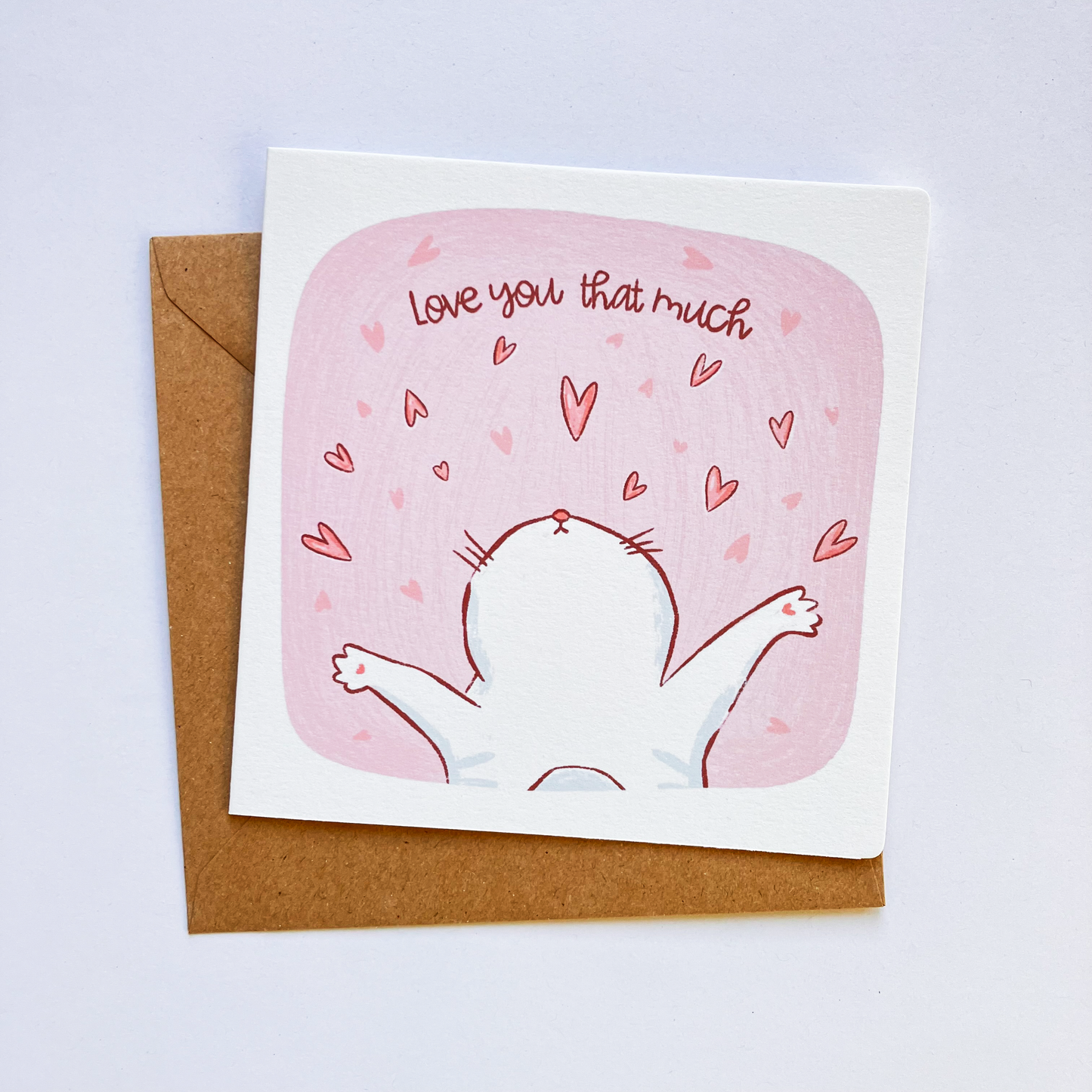 A greeting card with a whimsical illustration of a cat with paws spread, with the text 'Love you that much' above it. The card is placed on a white background with a brown envelope behind it