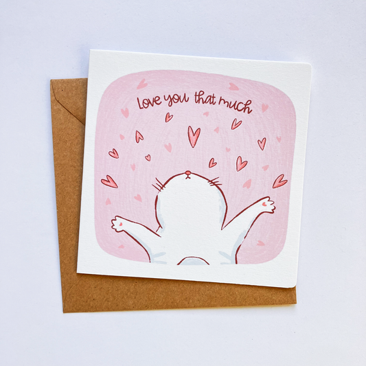 A greeting card with a whimsical illustration of a cat with paws spread, with the text 'Love you that much' above it. The card is placed on a white background with a brown envelope behind it
