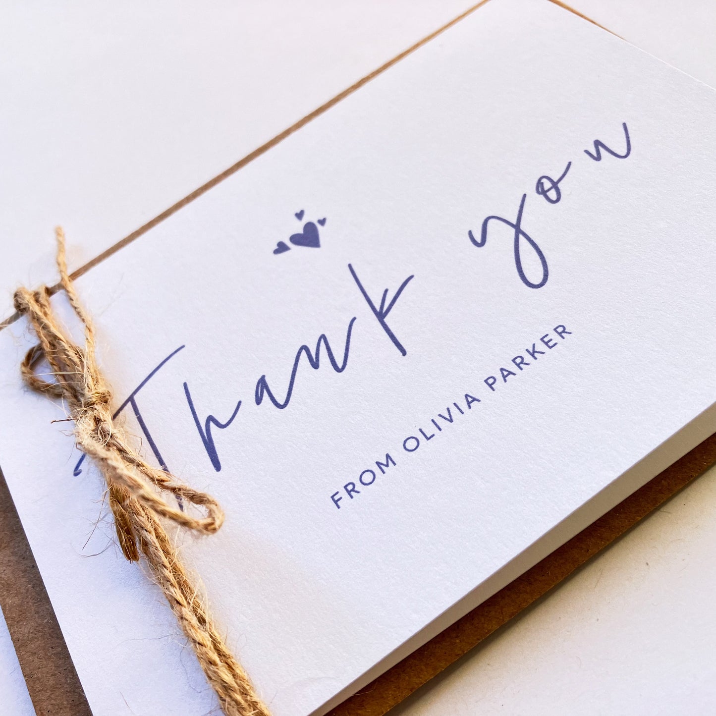 Angle view of a personalised thank you card with a handwritten "Thank you" text in the middle. On top of the text, there are four heart shapes, one big and three small ones on the left, right and top. Below the "Thank you" text is the personalisation with example one shown as "FROM OLIVIA PARKER". The card's background is soft lavender one and the text and illustrations are in dark muted purple. The card is wrapped on the left side with a natural baker thick thread.