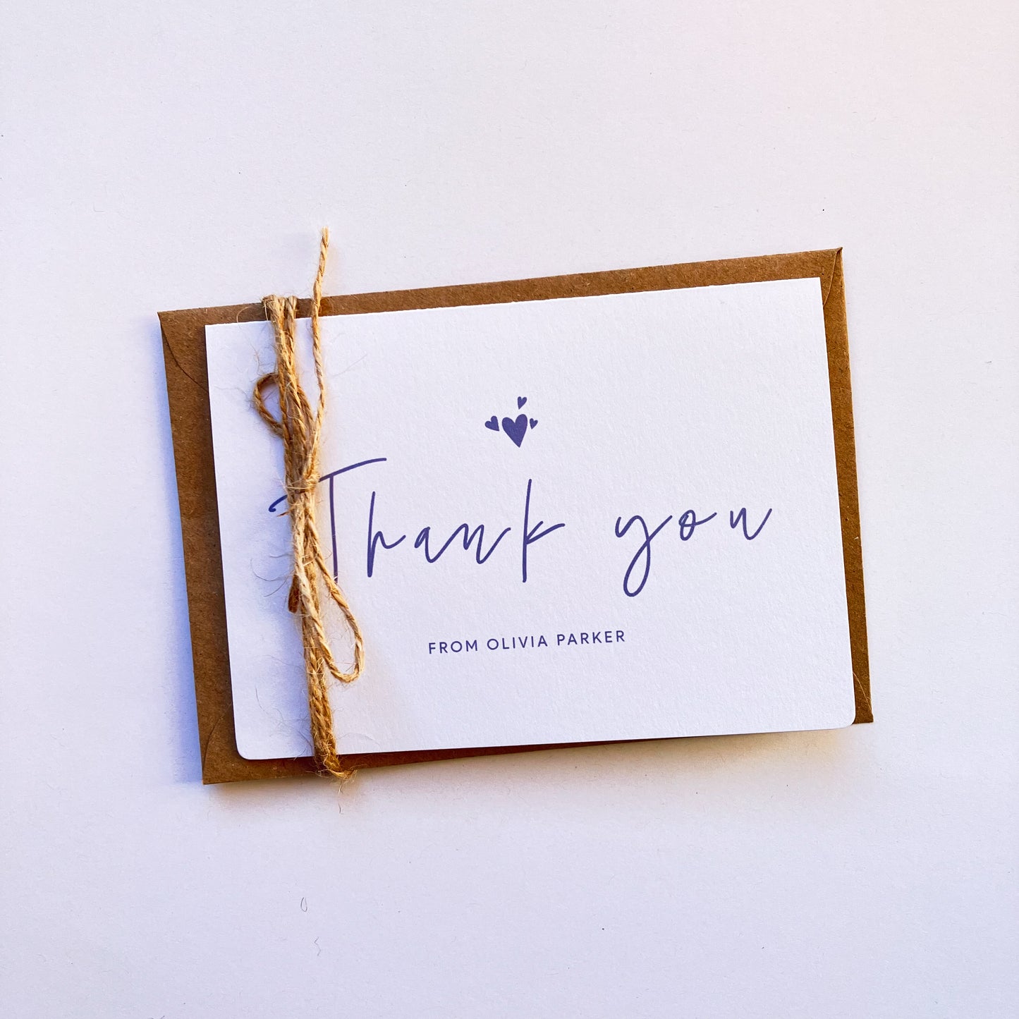 Top view of a personalised thank you card with a handwritten "Thank you" text in the middle. On top of the text, there are four heart shapes, one big and three small ones on the left, right and top. Below the "Thank you" text is the personalisation with example one shown as "FROM OLIVIA PARKER". The card's background is soft lavender one and the text and illustrations are in dark muted purple.