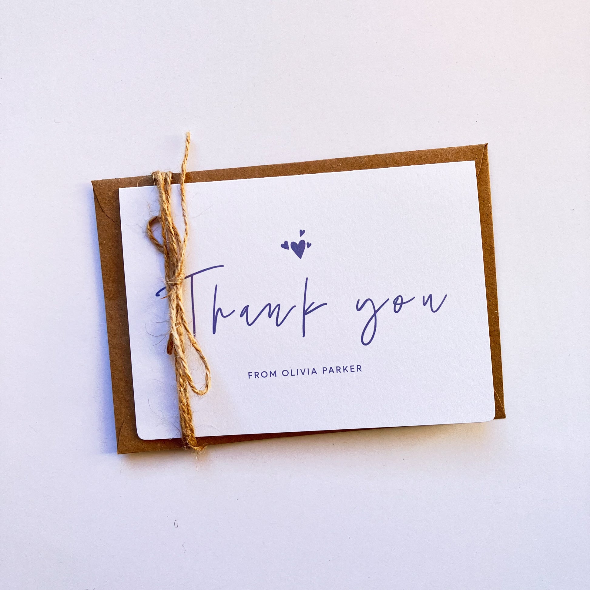 Top view of a personalised thank you card with a handwritten "Thank you" text in the middle. On top of the text, there are four heart shapes, one big and three small ones on the left, right and top. Below the "Thank you" text is the personalisation with example one shown as "FROM OLIVIA PARKER". The card's background is soft lavender one and the text and illustrations are in dark muted purple.