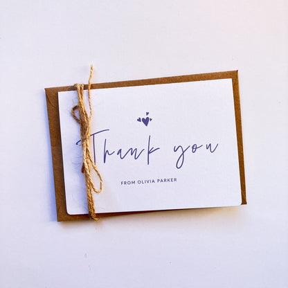 Top view of a personalised thank you card with a handwritten "Thank you" text in the middle. On top of the text, there are four heart shapes, one big and three small ones on the left, right and top. Below the "Thank you" text is the personalisation with example one shown as "FROM OLIVIA PARKER". The card's background is soft lavender one and the text and illustrations are in dark muted purple.