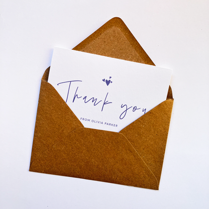 Top view of a personalised thank you card with a handwritten "Thank you" text in the middle. On top of the text, there are four heart shapes, one big and three small ones on the left, right and top. Below the "Thank you" text is the personalisation with example one shown as "FROM OLIVIA PARKER". The card's background is soft lavender one and the text and illustrations are in dark muted purple. The card is inside a kraft brown envelope.