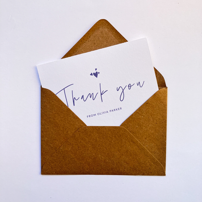 Top view of a personalised thank you card with a handwritten "Thank you" text in the middle. On top of the text, there are four heart shapes, one big and three small ones on the left, right and top. Below the "Thank you" text is the personalisation with example one shown as "FROM OLIVIA PARKER". The card's background is soft lavender one and the text and illustrations are in dark muted purple. The card is inside a kraft brown envelope.