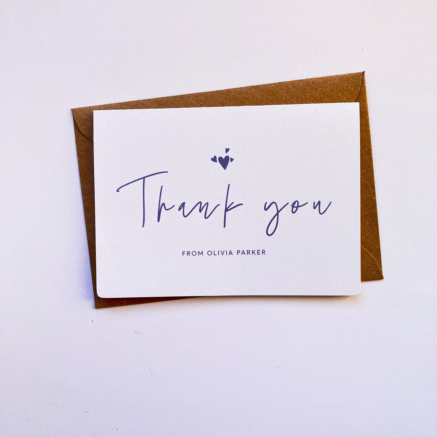 Top view of a personalised thank you card with a handwritten "Thank you" text in the middle. On top of the text, there are four heart shapes, one big and three small ones on the left, right and top. Below the "Thank you" text is the personalisation with example one shown as "FROM OLIVIA PARKER". The card's background is soft lavender one and the text and illustrations are in dark muted purple. The card is on top of a kraft brown envelope.
