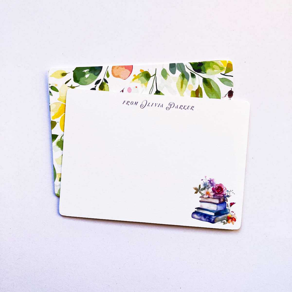 Custom notecard with floral book art and personalised text, displayed on a closed kraft envelope with vibrant yellow watercolour floral cards behind it.