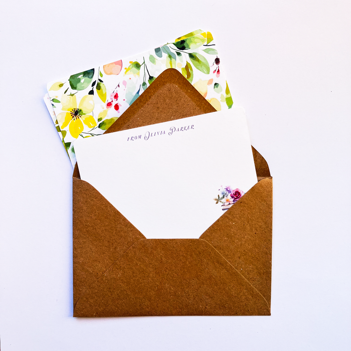 Open brown kraft envelope revealing a personalised notecard with a watercolour book illustration, accompanied by matching bright floral stationery in the background